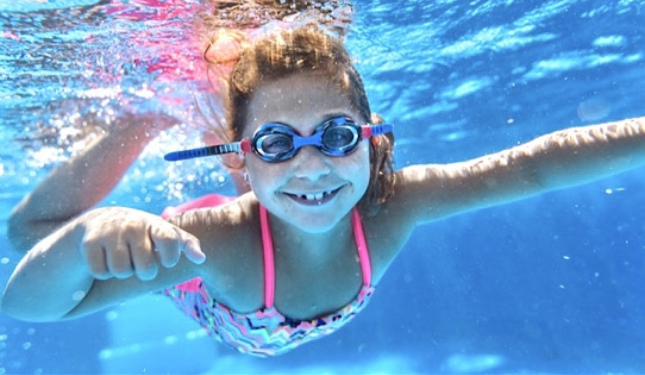 Mädchen unter Wasser mit Schwimmbrille lächelt ins Bild 