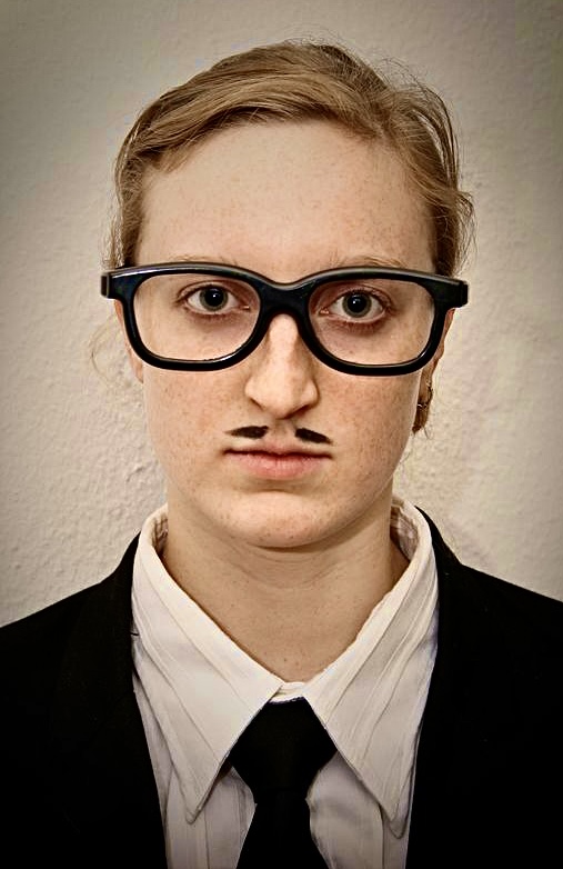 Junge Frau mit Anzug, schwarzer Brille und Schnurrbart blickt ins Bild