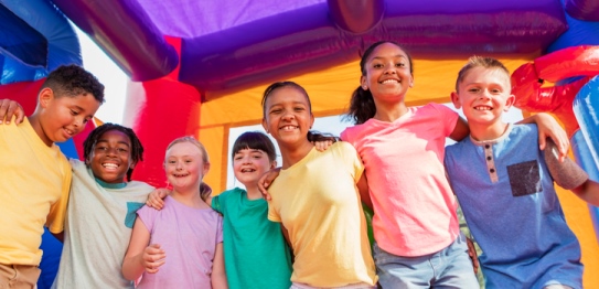 Eine Reihe von lachenden multiethnischen Kindern, die sich die Arme auf die schultern gelegt haben in einem bunten H&uuml;pfhaus  