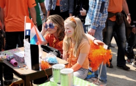 Zwei Mädchen mit orangenen T-shirts und Sonnenbrille sitzen am Flohmarktisch, Menschenmenge im Hintergrund